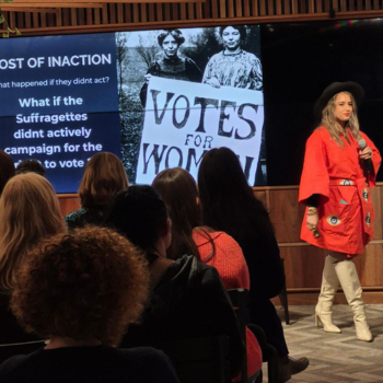 Speaker in red jacket presenting to an audience with a “Votes for Women” slide behind.